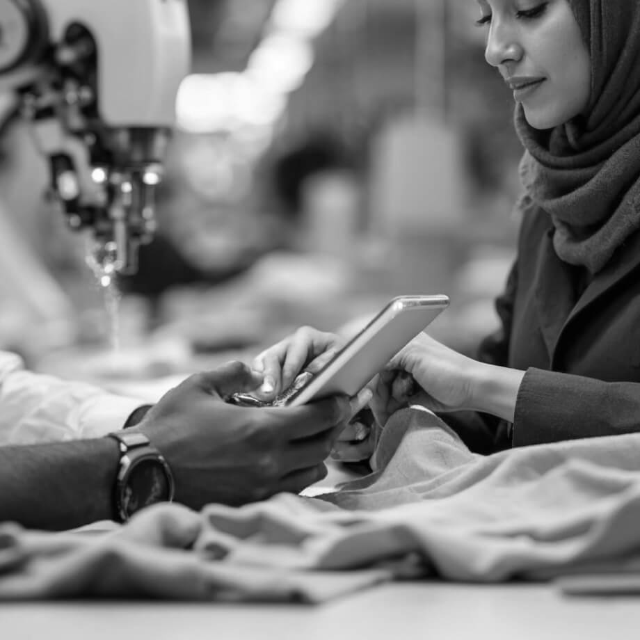 Two people work together at a sewing station. One operates a smartphone, and the other helps adjust fabric near a sewing machine. The environment suggests a garment production setting. The image is in black and white.